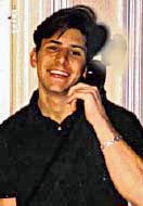 A young Caucasian man with dark hair, wearing a dark collared shirt with the top button opened, smiling and holding a telephone handset to his left ear. Behind him is a striped background; there is a pixillated area to the top right of his head.