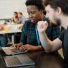 A white male employee follows along as a Black female employee walks him through work on her laptop.