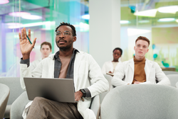 Front view portrait of African American man wearing lab coat and raising hand asking question while sitting in audience and listening to lecture on medicine