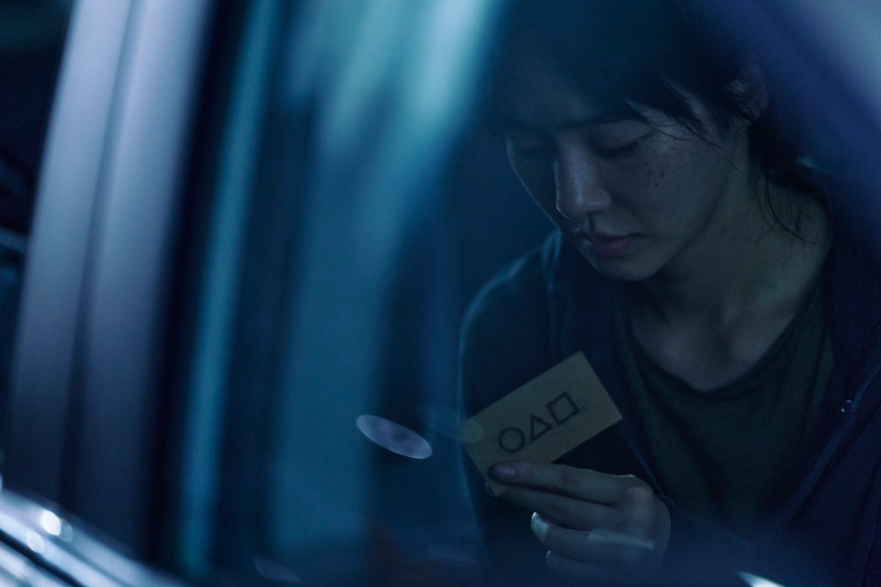 A woman sits in a car reading a small card with a circle, triangle, and square on it in 'Squid Game' Season 2 