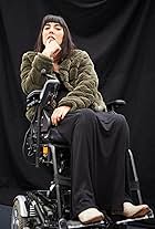 Kyla, a person of colour in her 30’s with dark hair, wearing a green fake fur coat, sits in a raised power chair, looking defiantly into the camera.