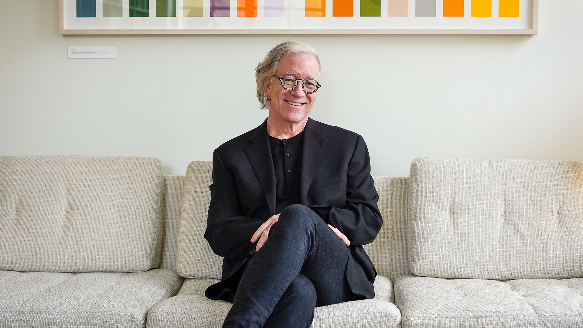 Jeff MacKie-Mason, dressed in all black, sits on a couch and smiles for a portrait