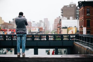 A man stands on a bridge, photographing a vibrant cityscape, suggesting a neurodivergent or different way of looking at the world.