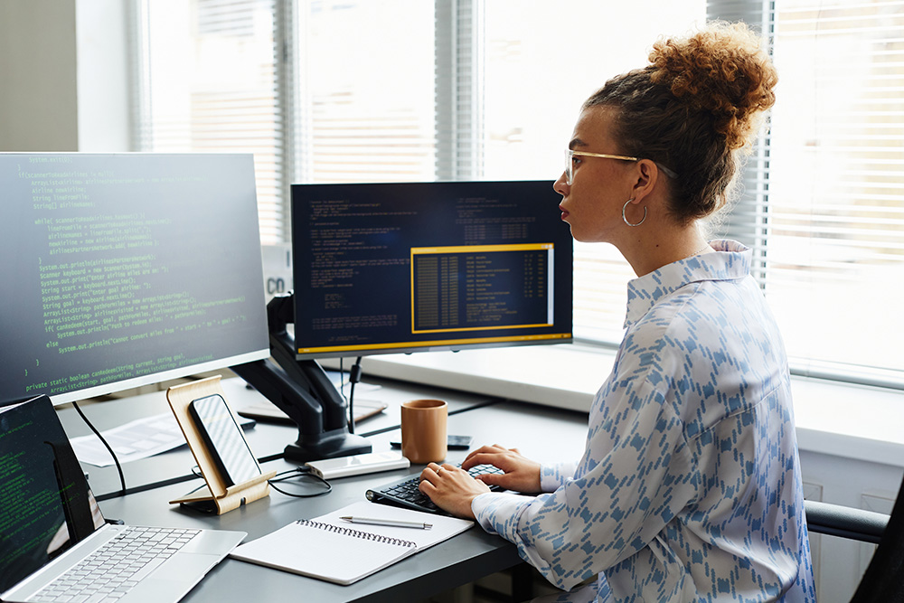 Eine junge Frau sitzt an einem Schreibtisch vor zwei Computer-Monitoren und programmiert.